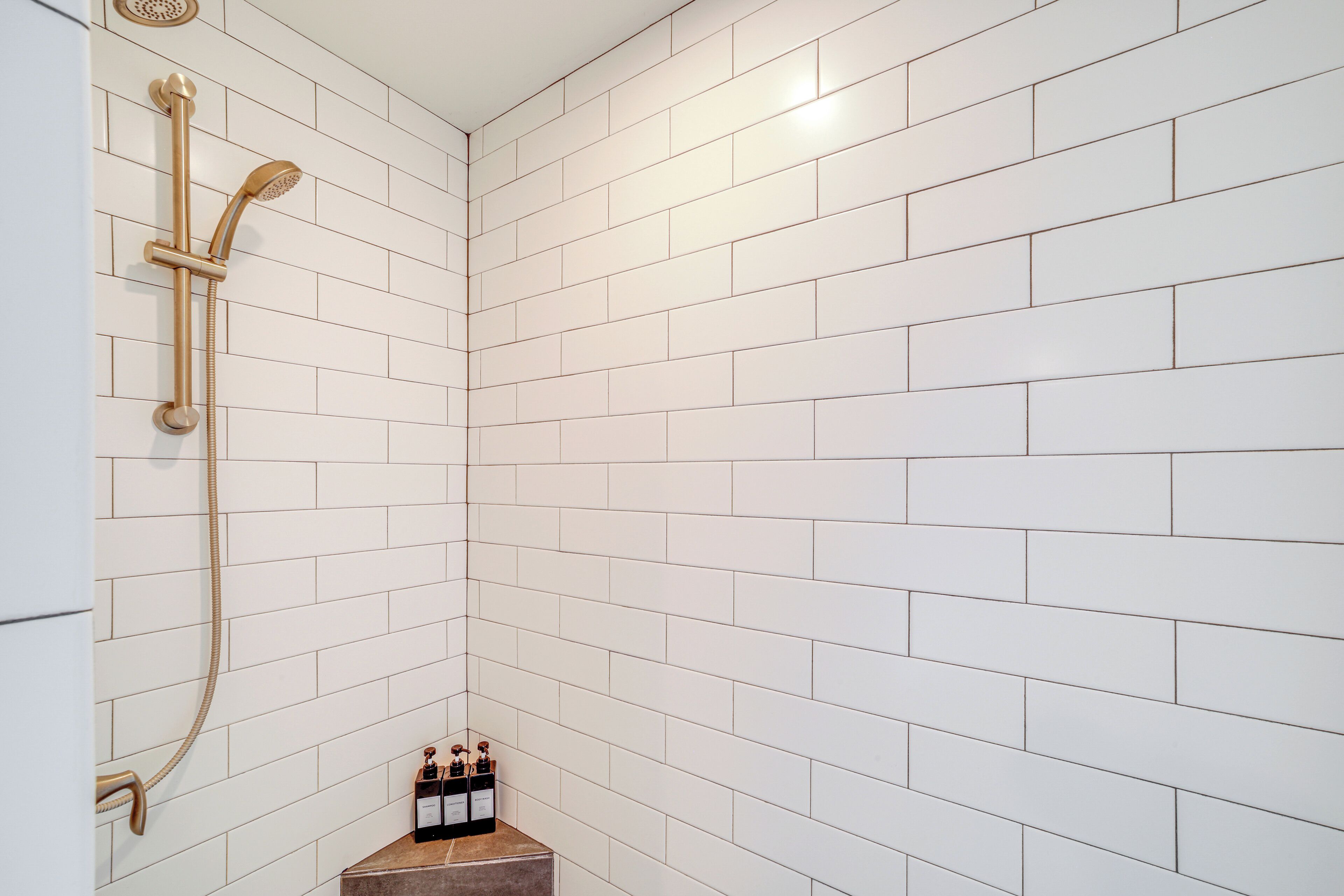 Une salle de bain avec des carreaux de céramique blancs rectangulaires, une pomme de douche dorée et une bouteille de shampoing posée sur une petite étagère.