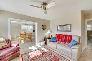 Een woonkamer met een beige bank, een tapijt met rood-wit patroon, een plafondventilator, een eethoek met tafel en stoelen, en een klok aan de muur.