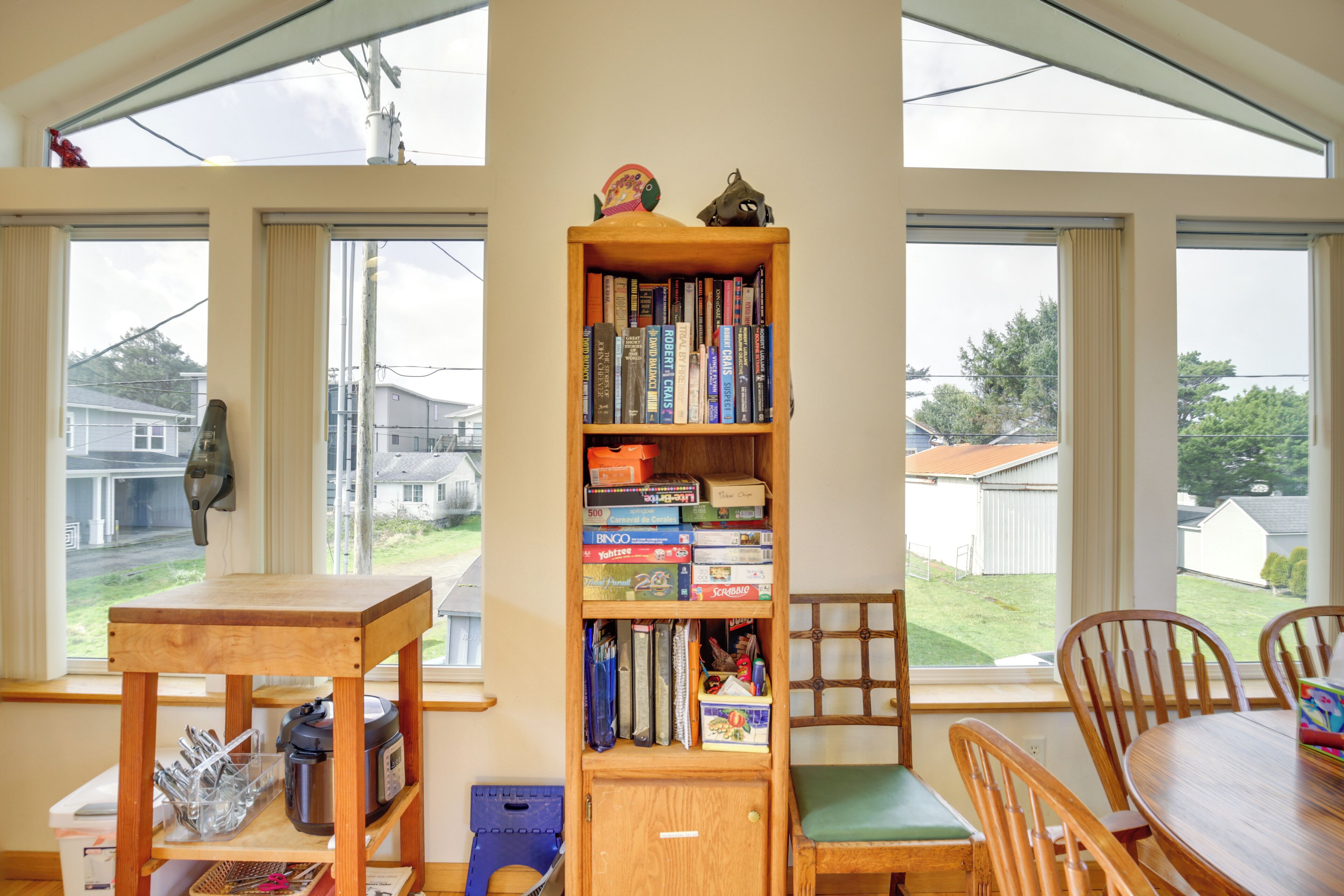 A room with a wooden bookshelf filled with books, a dining table with chairs, and large windows offering a view of the outside.