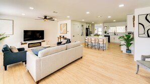 A modern living room with a sofa, TV, and ceiling fan. A kitchen with bar stools is visible in the background.