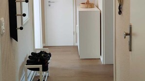 A hallway with a wooden floor, a white door at the end, a small white cabinet, and a shoe rack with shoes.
