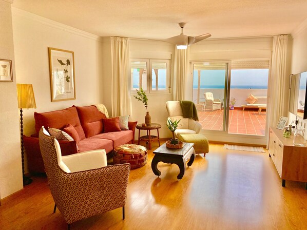 A living room with a sofa, armchair, and a coffee table. There is a ceiling fan, a window with curtains, and a view of the sea.