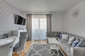 Un salon moderne avec un canapé gris, une table basse blanche et une table à manger grise assortie à ses chaises. Un téléviseur à écran plat est fixé au mur, et une horloge décorative est visible.