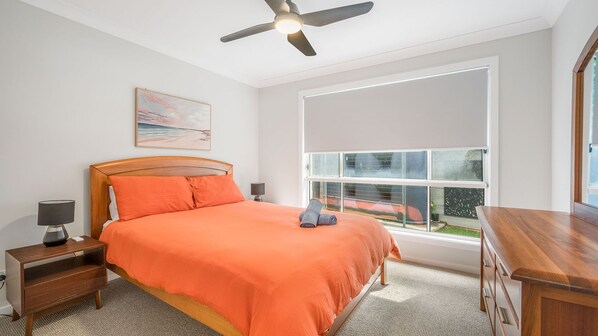 Une chambre à coucher comprenant un lit, une table de chevet, un bureau, un ventilateur de plafond, une fenêtre et un tableau accroché au mur.