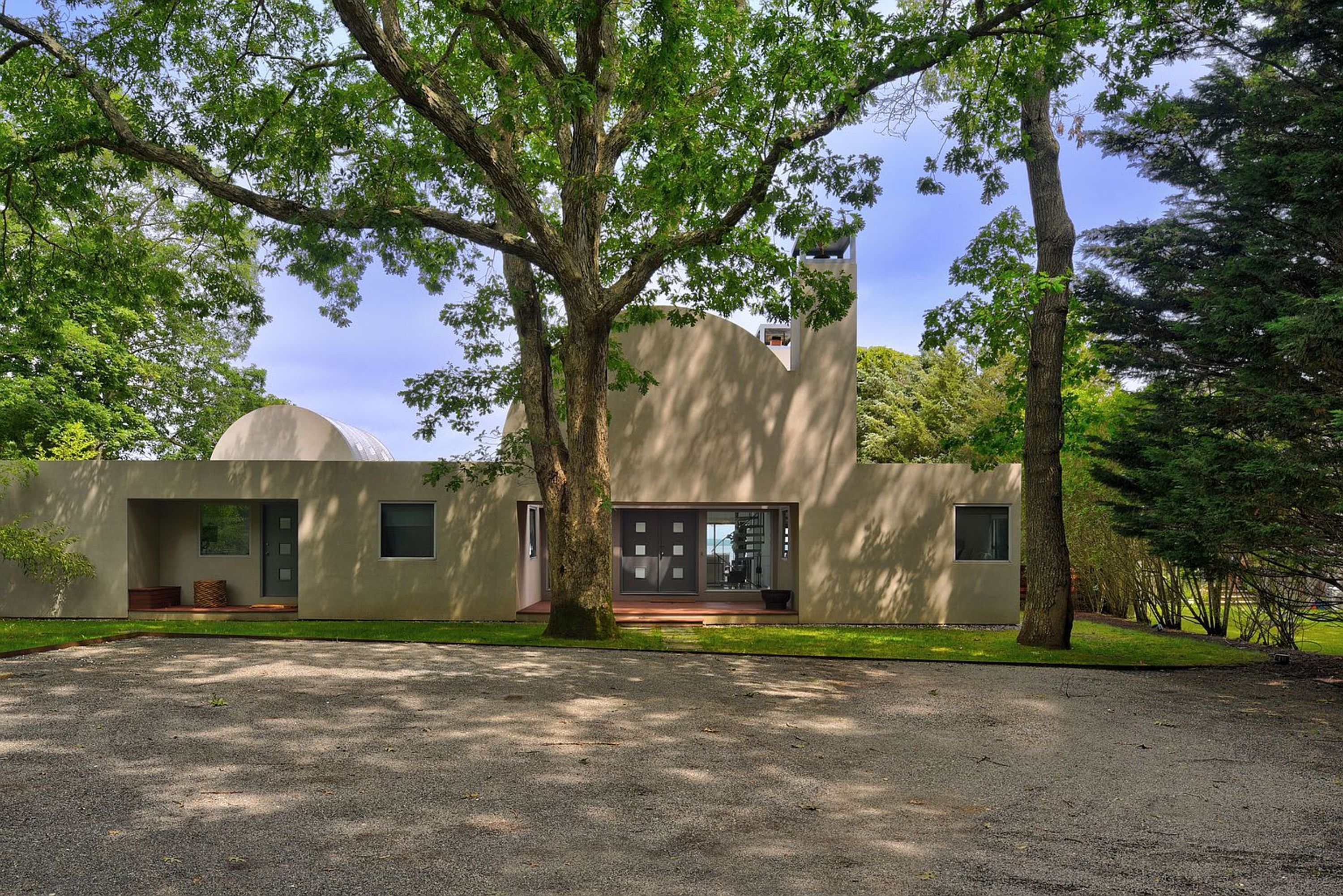 A modern building with a large tree in front, a gravel driveway, and a clear sky.