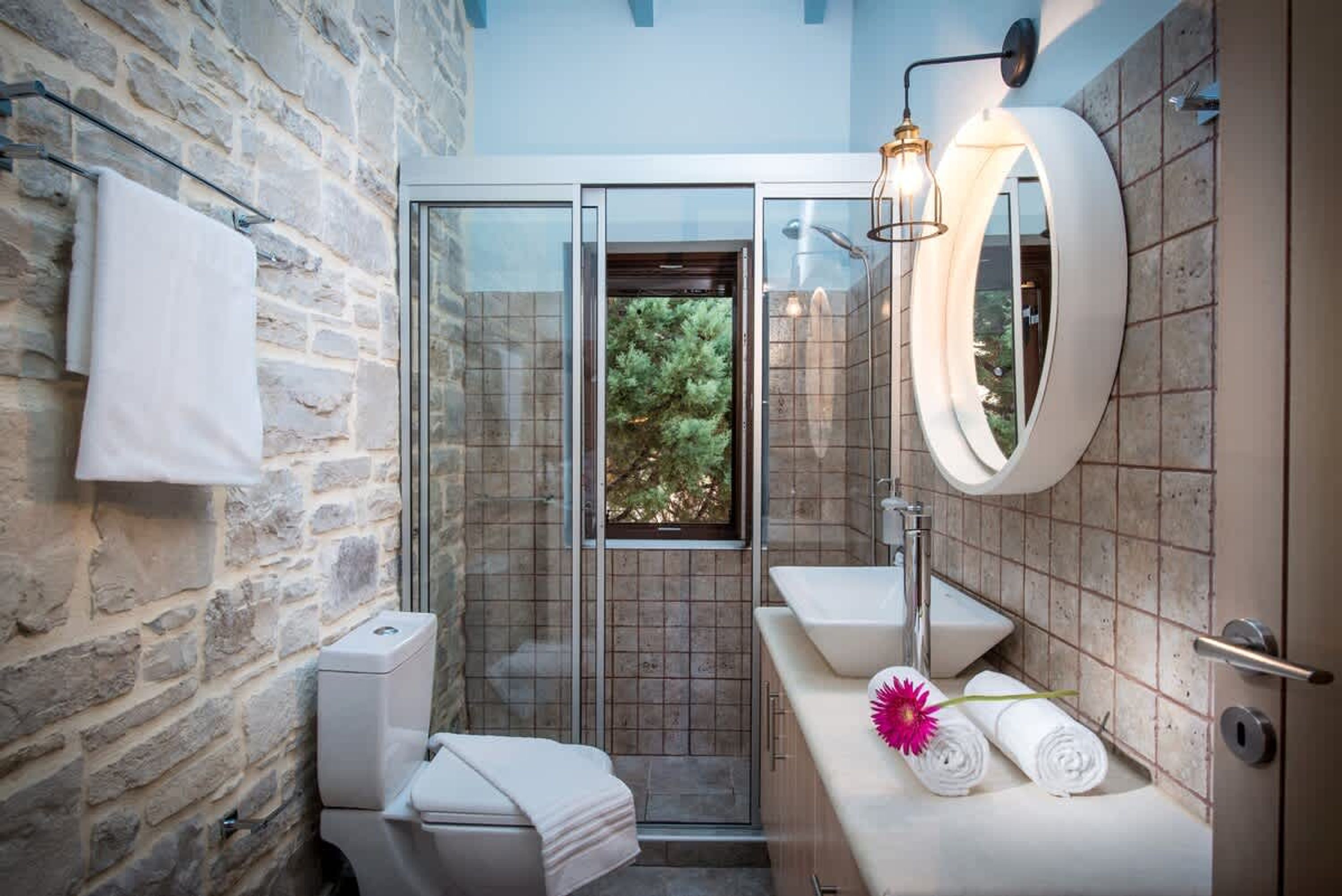 Un baño con pared de piedra, cabina de ducha de vidrio, lavamanos blanco y espejo.