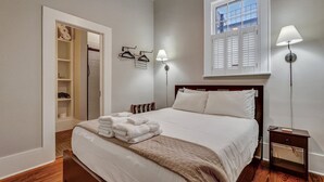 Una cama bien hecha con una colcha blanca y una manta marrón, una mesita de noche con una lámpara, y una ventana con contraventanas blancas.