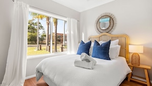 A hotel room with a large bed, white bedding, a wooden headboard, a bedside lamp, and a view of palm trees and a building outside.