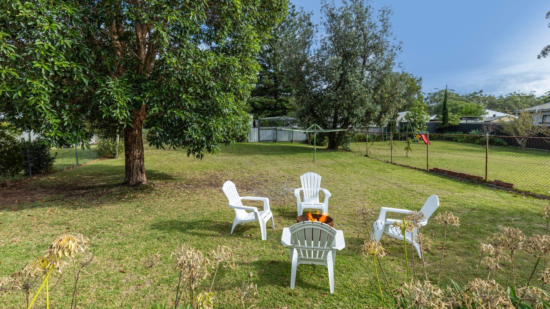 Un espace extérieur verdoyant, avec des chaises en plastique blanches disposées en demi-cercle, un arbre et, au fond, une aire de jeux clôturée.
