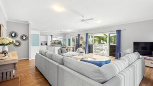 A modern living room with a grey sectional sofa, a coffee table, and a TV. There is a kitchen in the background with white cabinets and a dining area with a table and chairs.