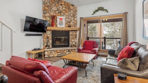 A cozy living room with a stone fireplace, leather sofas, a flat-screen TV, and a large window overlooking a deck. - Cozy Top Floor Unit, Near Main St. Hot tubs & Pool (Breckenridge)