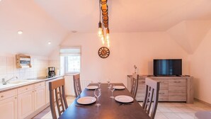 Een eetgedeelte met een lange houten tafel, gedekt voor vier personen, een klok aan de muur en een televisie op een kast.