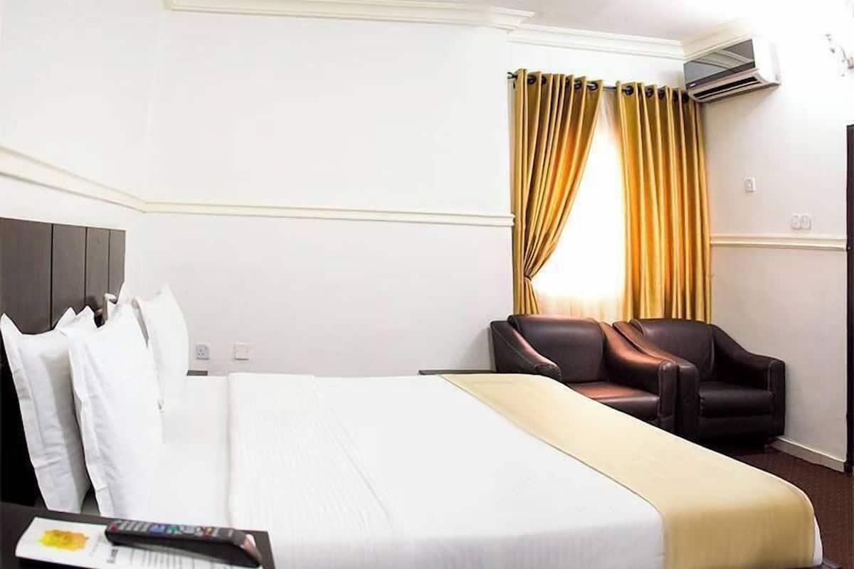 A hotel room with a bed, a desk with a remote control, a leather chair, and a window with curtains.