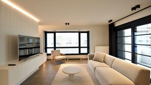 A modern living room with a flat-screen TV, a beige sofa, a round coffee table, and a large window providing a view of the city. - Amazing Apartment in De Haan Near Beach (De Haan)
