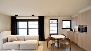 A modern living room with a beige sofa, a round dining table with chairs, a kitchen area with wooden cabinets, and large windows with black curtains. - Amazing Apartment in De Haan Near Beach (De Haan)