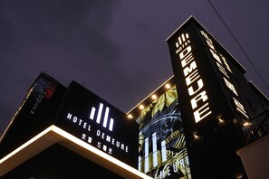 Ein Hotel mit beleuchteter Beschilderung, darunter Schriftzüge mit "HOTEL DEMEURE" und "HOTEL DE MEURE" in großen Lettern.