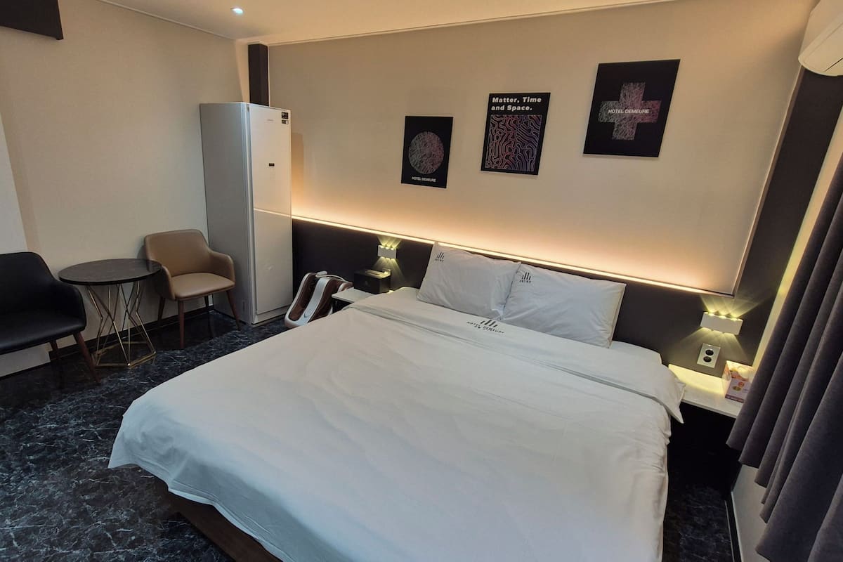 A hotel room with a bed, a chair, a small table, a minibar, and wall art.
