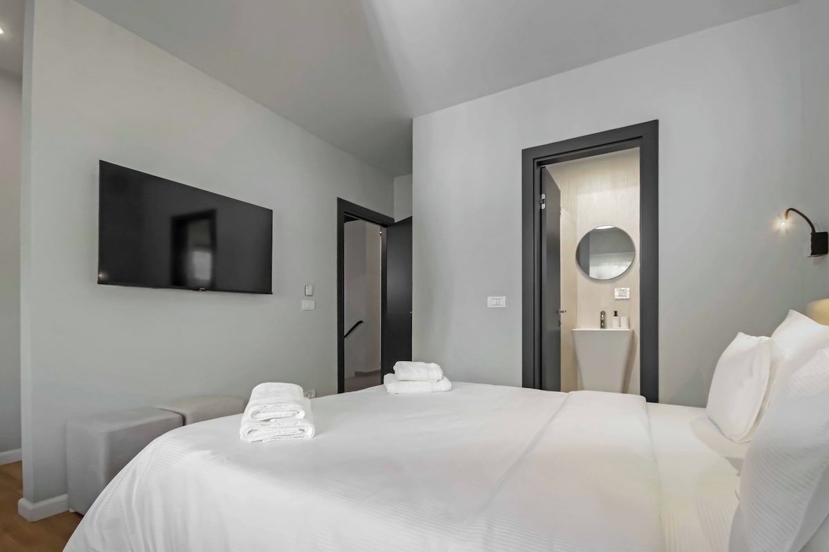 Habitació d'hotel moderna amb un llit gran, una televisió de pantalla plana penjada a la paret, una tauleta de nit amb una làmpada i una porta que dona a un bany amb mirall i rentamig.