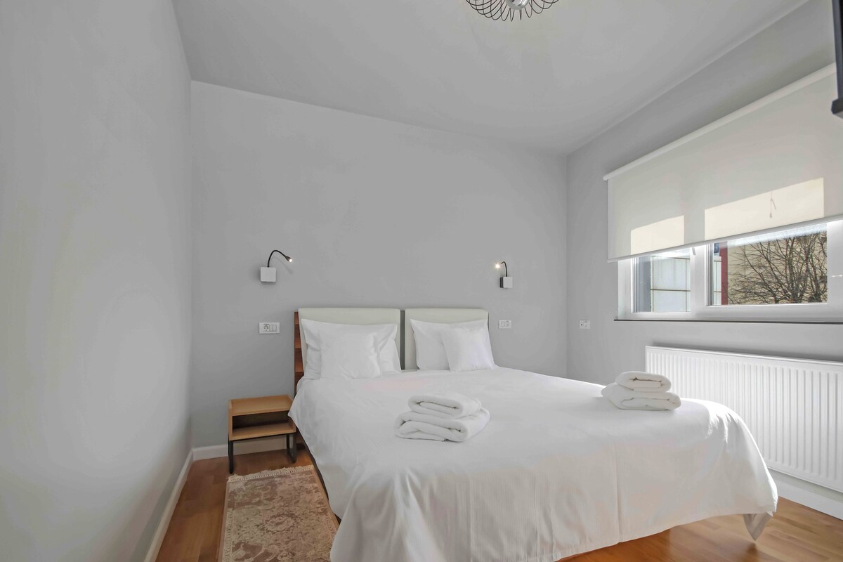 A bedroom with a large bed, two bedside tables, a bench, and a window with a view.