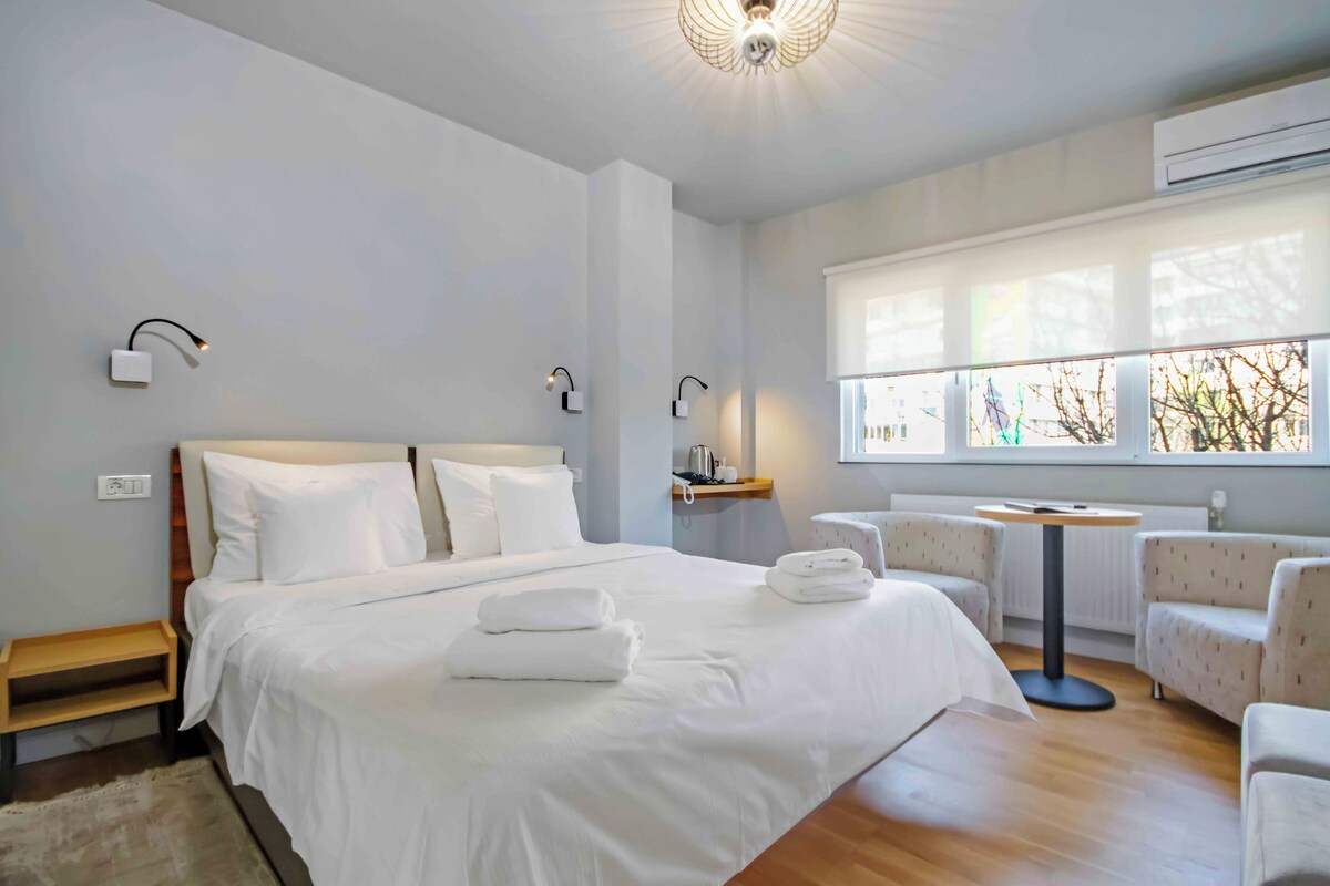 Ein modernes Hotelzimmer mit einem großen Bett, Nachttischen, einem kleinen runden Tisch und einem Fenster mit Blick auf Bäume.