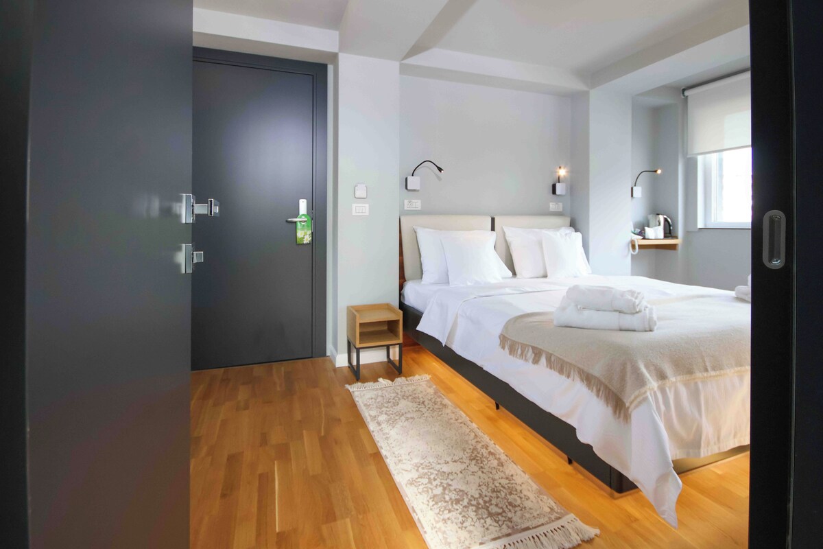 A modern bedroom with a large bed, a wooden floor, a dark door, and a small bedside table.