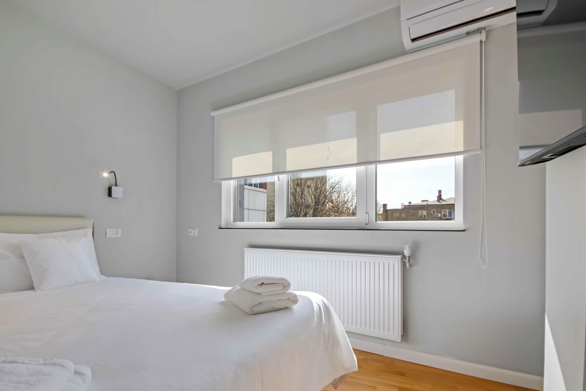A modern hotel room with a neatly made bed, a window with blinds, a radiator, and a view of buildings outside.