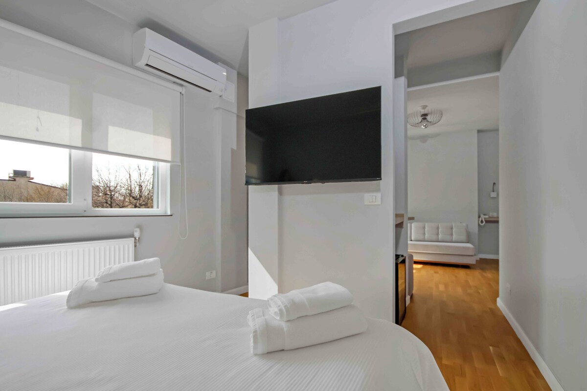 A modern hotel room with a large bed, a flat-screen TV mounted on the wall, and a view of the city through the window.