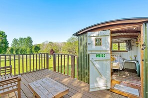 A wooden deck with a bench and a table, a small room with a bed and a window, and a green sign indicating a first-floor room. - Couples Rural Retreat – Shepherd’s Hut at Hilltop (Romsey)