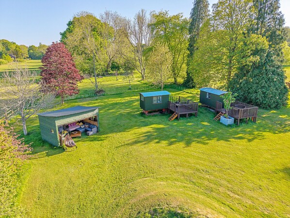 A campsite with two green cabins, a small shed, and a deck area with seating. - Couples Rural Retreat – Shepherd’s Hut at Hilltop (Romsey)