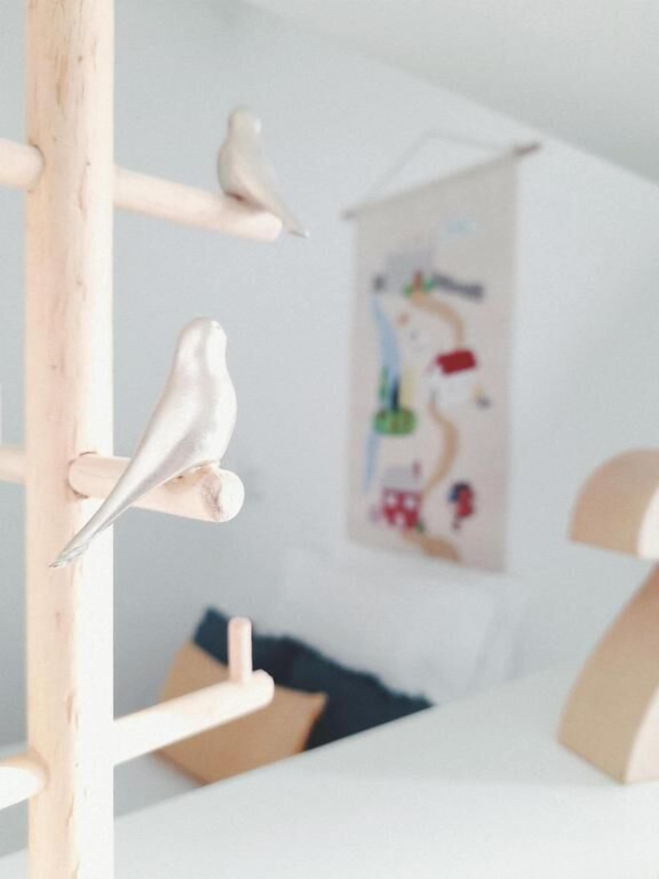 Een kamer met een houten constructie, mogelijk een plank of meubelstuk, waarop twee witte vogelbeeldjes staan. Achtergrond is een kleurrijk abstract schilderij.