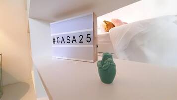 A bed with white bedding and a green decorative item on a white bedside table. A sign with "#CASA25" is visible on the wall.