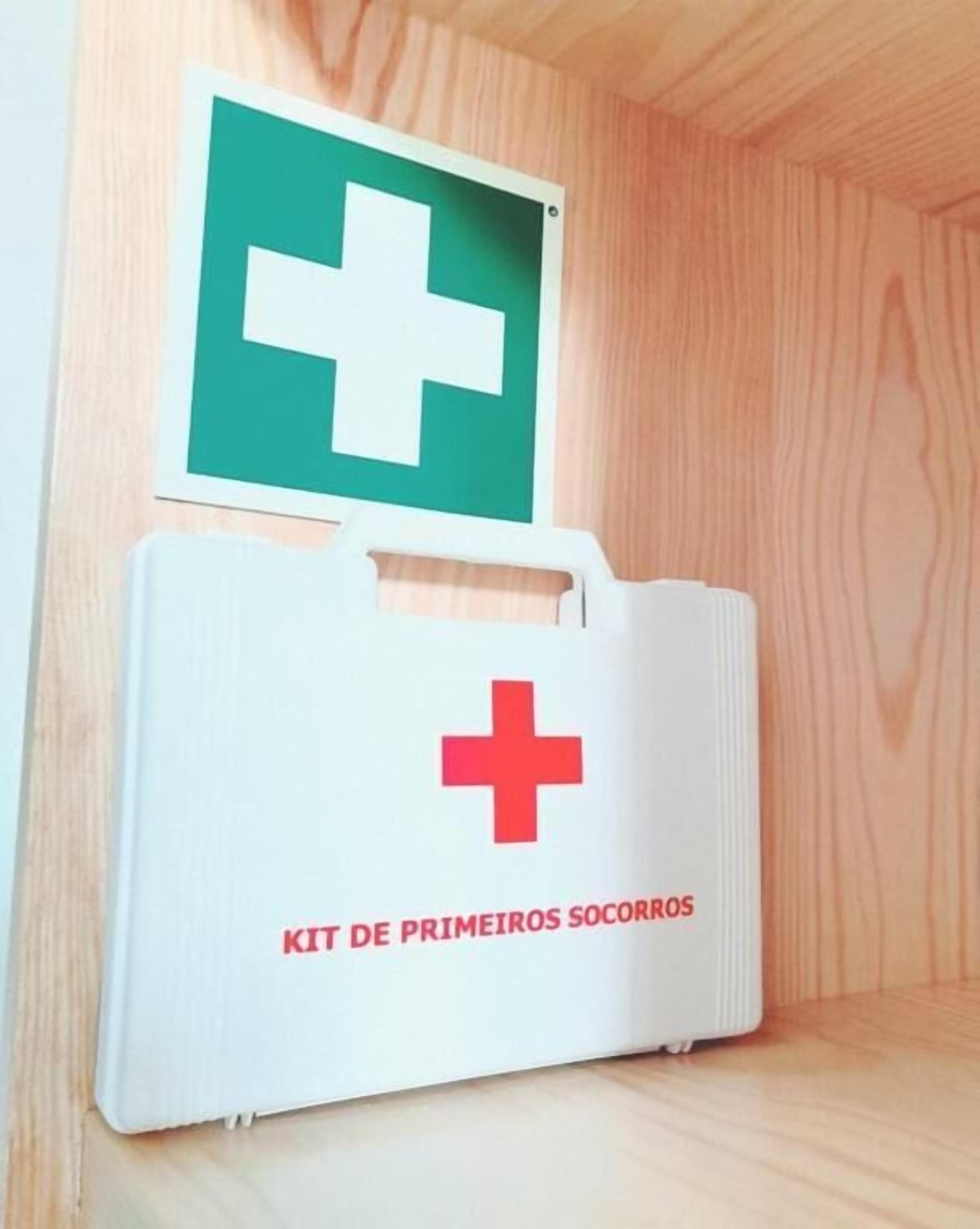 A wooden cabinet with a white first aid kit labeled "KIT DE PRIMEIROS SOCORROS" and a green emergency exit sign above it.