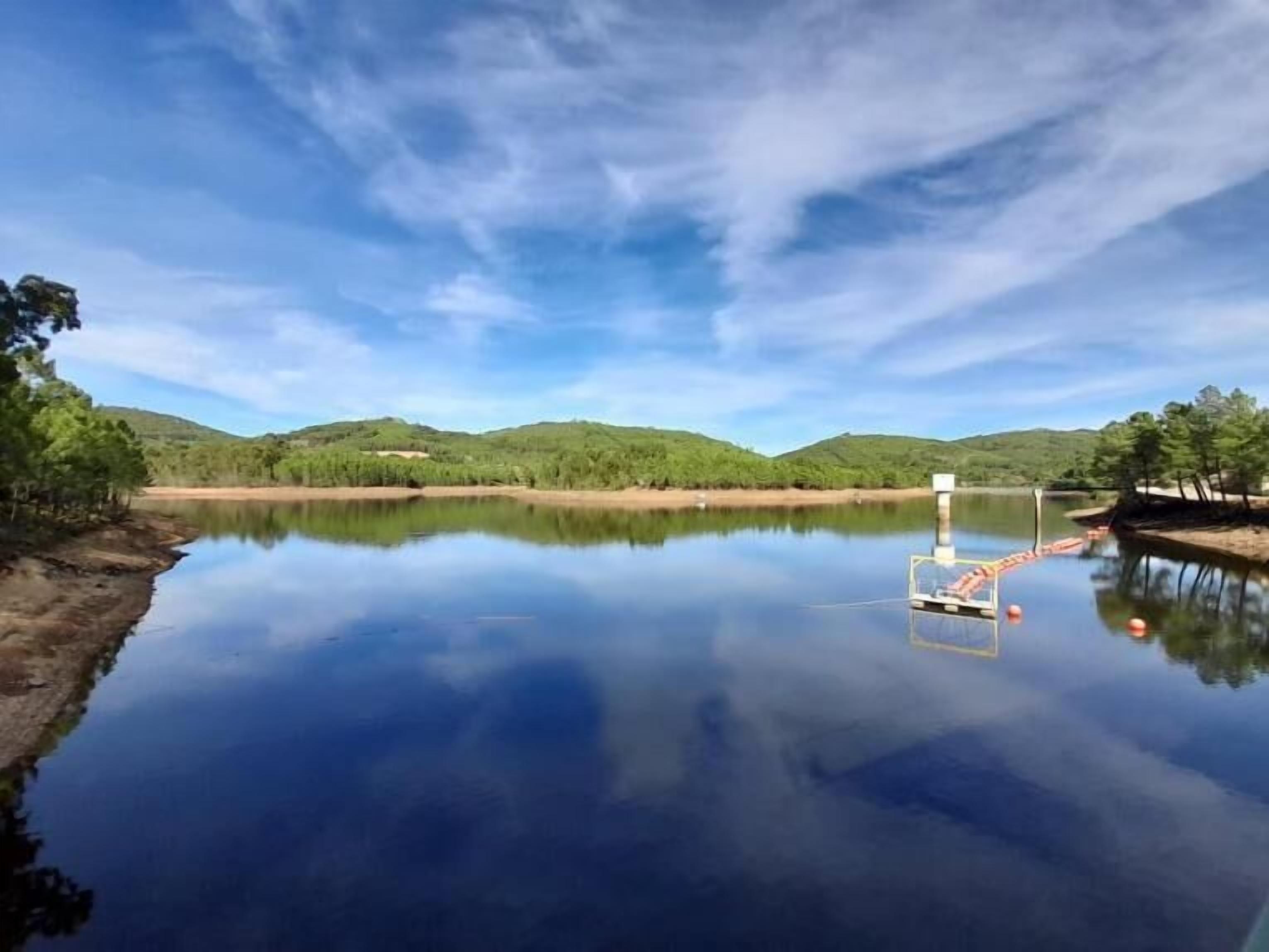 Een rustig meer met een steiger die het water in reikt, omringd door groene heuvels en bomen onder een heldere lucht.