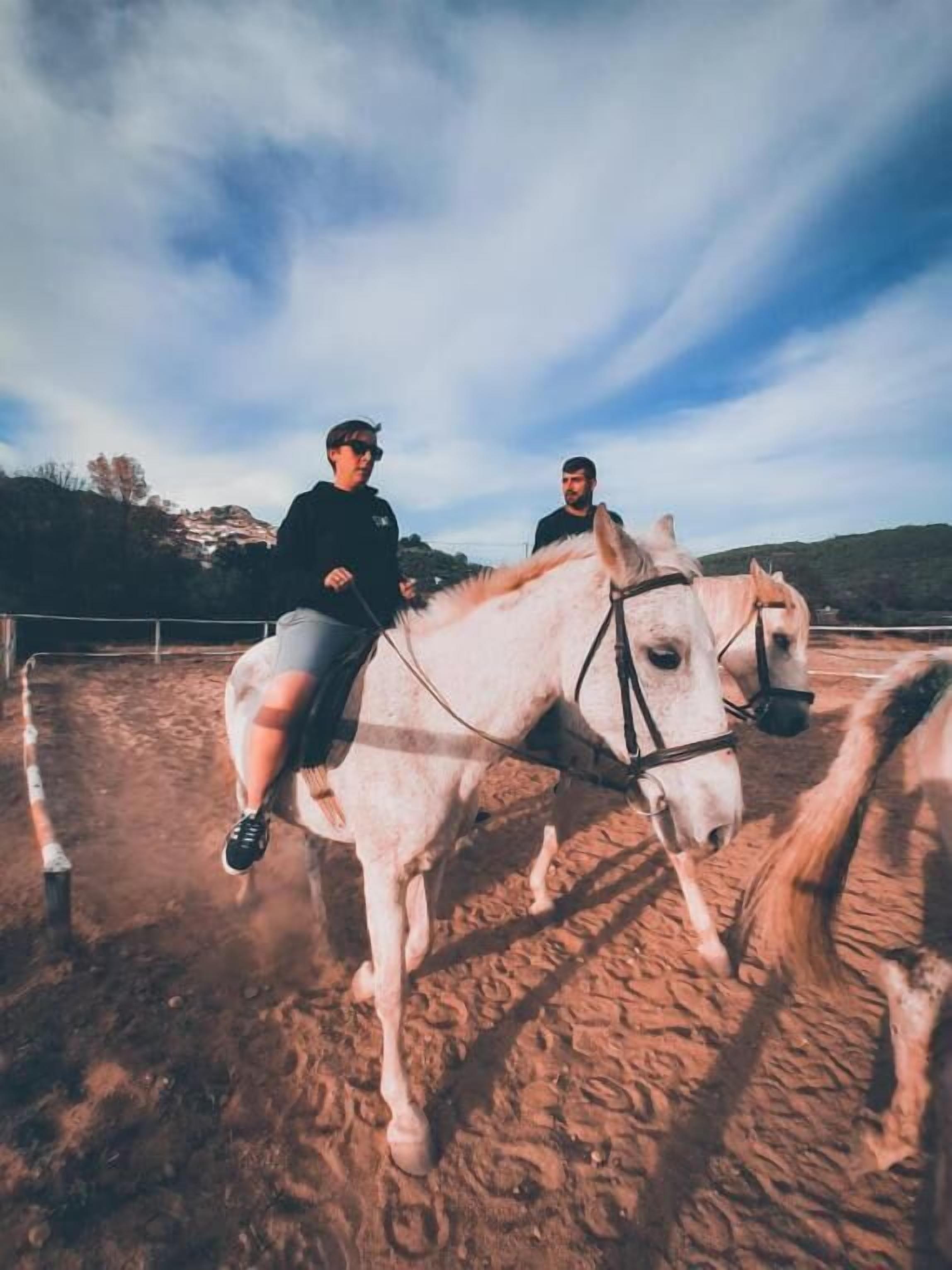 Dos personas montando a caballo en un picadero al aire libre con suelo de tierra y una valla de metal.