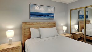 Une chambre d’hôtel avec un lit doté d’une tête de lit en bois, du linge de lit blanc, des lampes de chevet, un grand miroir et un tableau représentant une scène de plage.