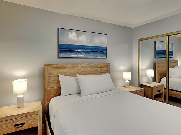 Ein Hotelzimmer mit einem Bett mit Holz-Kopfteil, weißer Bettwäsche, Nachttischlampen, einem großen Spiegel und einem Bild mit Strandmotiv.