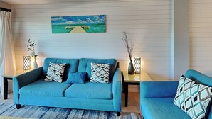 Ein Wohnzimmer mit einem blauen Sofa, einem Couchtisch und einem Gemälde eines Piers an der Wand.