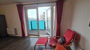 A room with a wooden floor, a red chair, a red cushion, and a view through a glass door to an outdoor balcony. - Barking 2 Bed Apt With Parking & Good Travel Links (Barking)