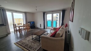 A living room with a sofa, a coffee table, and a TV. There is a balcony with a view of buildings and a pool. - Barking 2 Bed Apt With Parking & Good Travel Links (Barking)