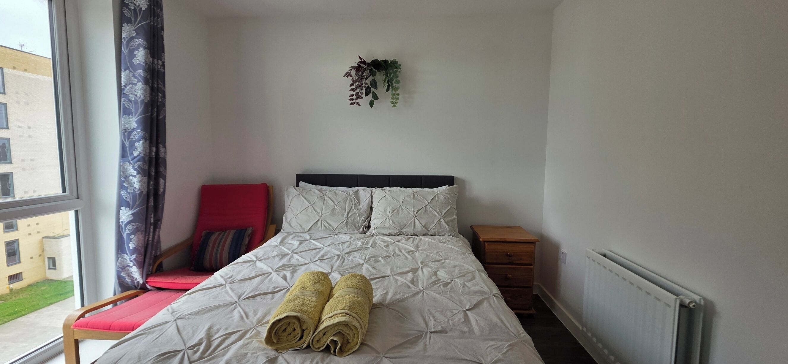 A neatly made bed with rolled towels, a red chair, a small bedside table, and a view of buildings outside the window.