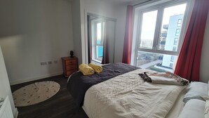 A hotel room with a large bed, a dresser, a mirror, and a view of the city through the window. - Barking 2 Bed Apt With Parking & Good Travel Links (Barking)