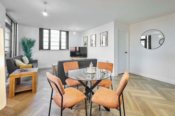 A modern living room with a glass dining table, orange chairs, a grey sofa, a TV, and a large mirror.