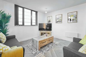 A modern living room with a flat-screen TV, a wooden coffee table, grey sofas, and a large window.
