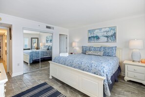 A bedroom with a large mirror, a bed with a blue patterned bedspread, a bedside table with a lamp, and a painting on the wall.