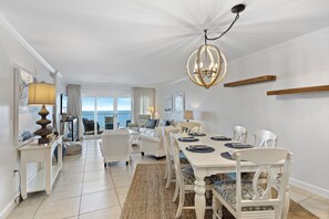 A spacious dining area with a white table set for six, surrounded by white chairs with patterned cushions. A large chandelier hangs from the ceiling, and the room features a view of the ocean through large windows.
