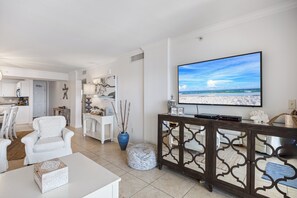 A living room with a large TV, white furniture, a decorative vase, and a view of the beach.