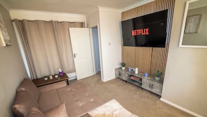 Olohuoneessa on ruskea sohva, seinälle asennettu televisio ja ovi, joka johtaa toiseen huoneeseen.
