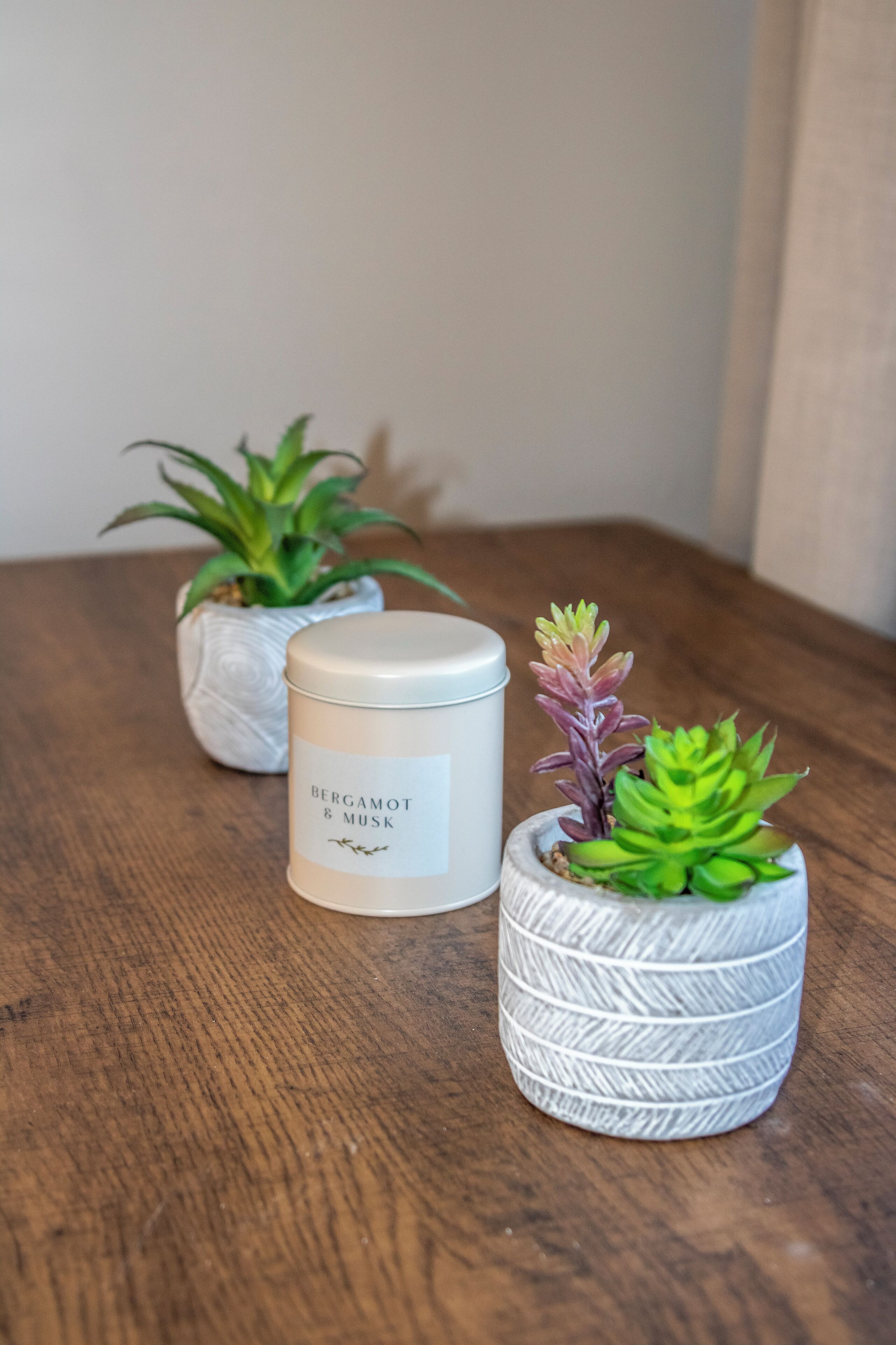 Un tavolo di legno con una candela bianca etichettata "BERGAMOT & MUSK" e due piccole piante in vaso.