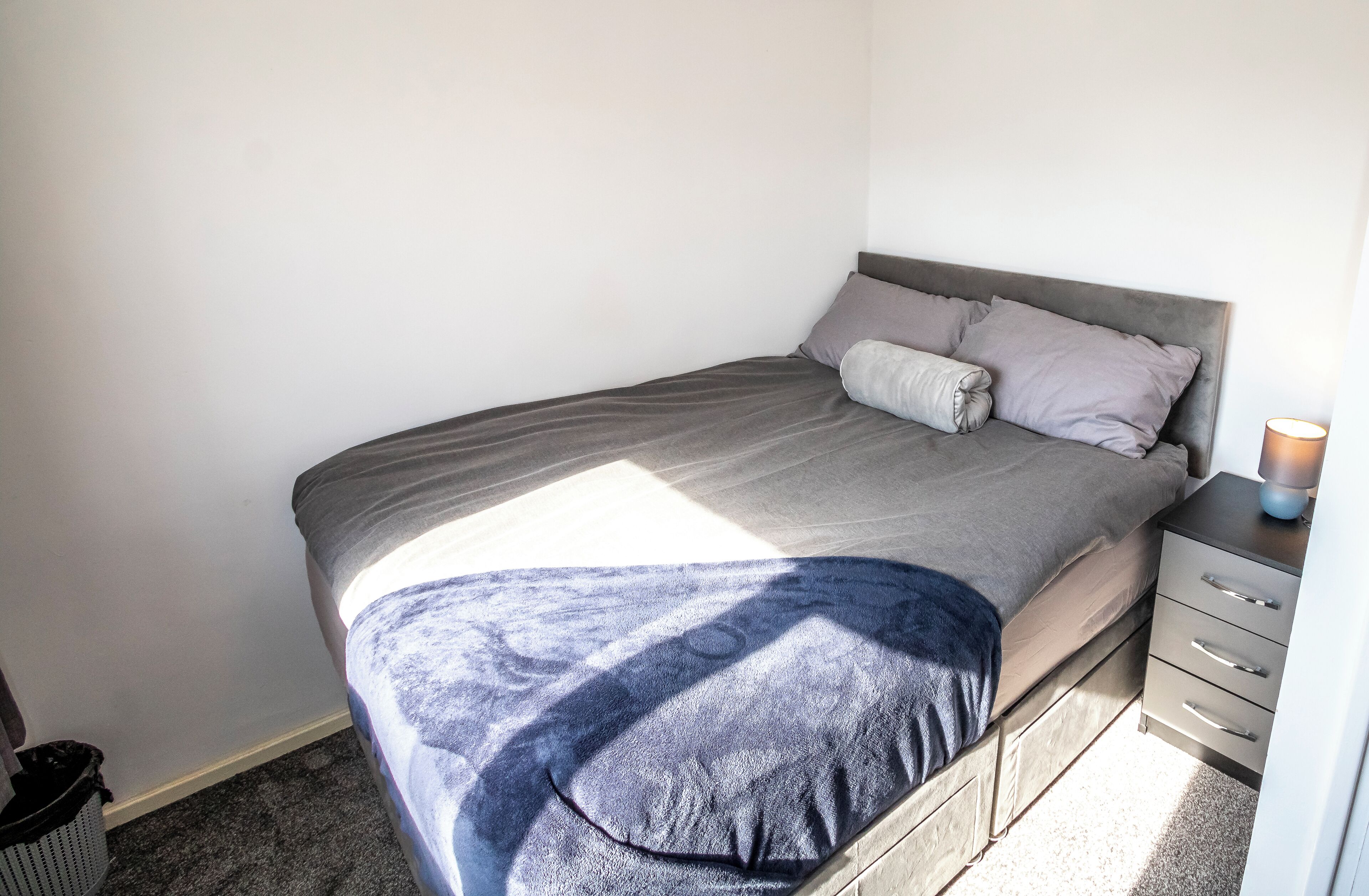 A neatly made bed with a grey headboard, a bedside table with a lamp, and a small trash bin.