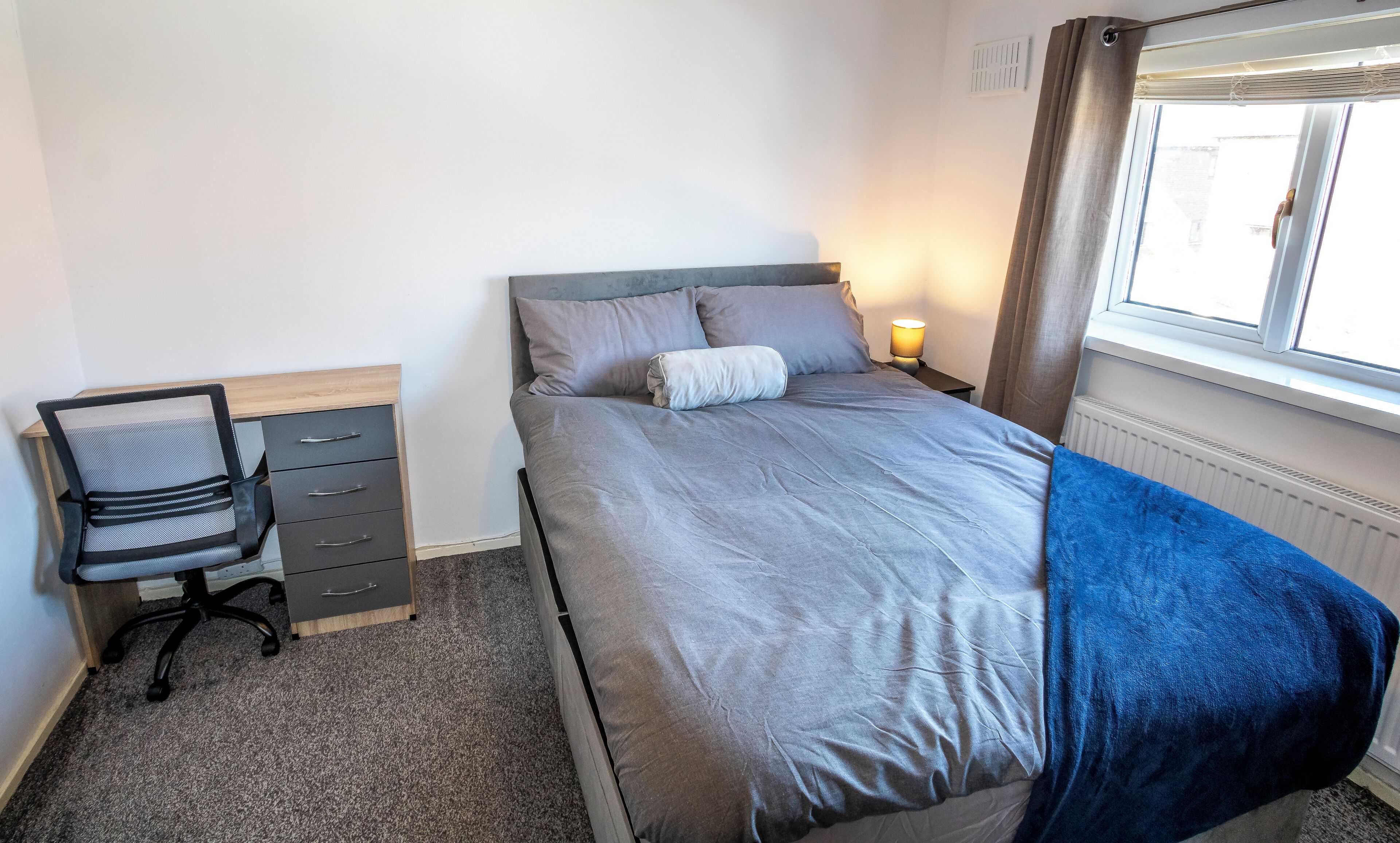 A bedroom with a bed, a desk with drawers, a chair, a lamp, and a window with curtains.